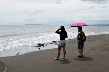 Bignay 2, Sariaya, Quezon, Philippines - December 3, 2020: Mature woman with hand bag and umbrella  and young lady sightseeing along sea shore