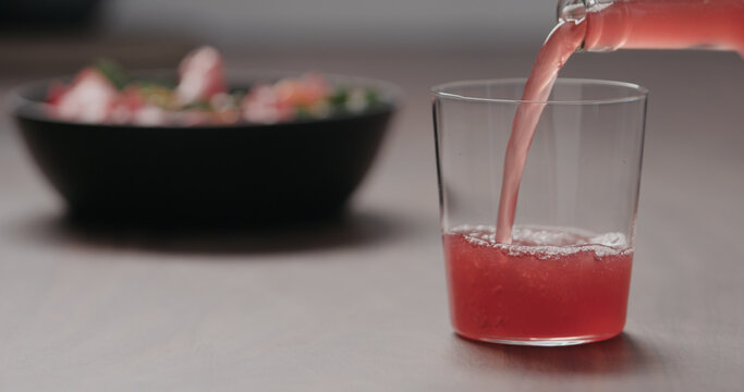 Pour Pink Fizzy Drink In A Tumbler Glass On Walnut Table With Salad On Background