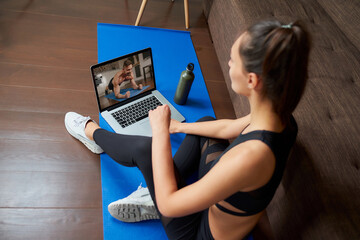 A laptop screen view over a woman's shoulder. A sporty girl in a tight suit is watching a workout video on laptop in her apartment. Woman is listening to a coach during a virtual fitness class at home