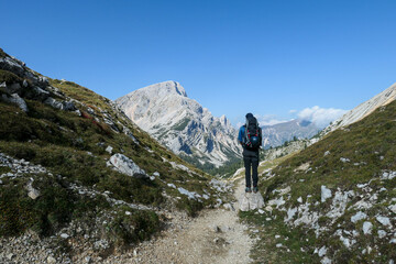 A man with a hiking backpack hiking on a narrow pathway in high Italian Dolomites. Steep, sharp mountain chain in the back. She is crossing a lush green meadow. Discovering and exploring the nature