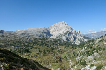 A panoramic view on a vast valley in Italian Dolomites. There are high mountain chains around. The bottom of the valley is lush green. A few trees on the slopes. Few soft clouds on the sky. Freedom