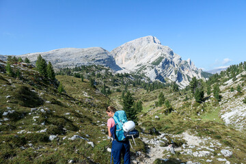 Naklejka na ściany i meble A woman with a hiking backpack hiking on a narrow pathway in high Italian Dolomites. Steep, sharp mountain chain in the back. She is crossing a lush green meadow. Discovering and exploring the nature