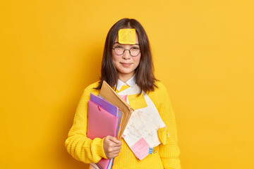 Pretty Asian female student with reminder sticker paper on forehead carries folders with papers prepares for difficult test wears round spectacles and jumper isolated over yellow background.