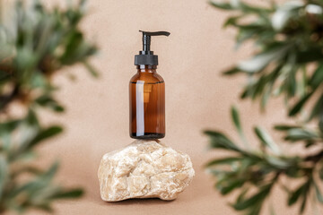 Brown glass bottle with pump of cosmetic products on stone framed by green leaves of branches, beige background. Natural Organic Spa Cosmetic Beauty Concept. Front view Mock up
