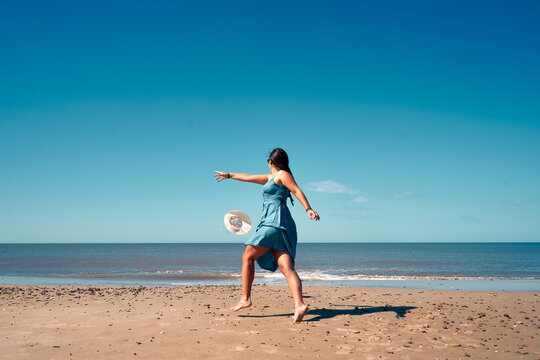 A backshot of a young female in a summer dress cathing up the hat on the beach