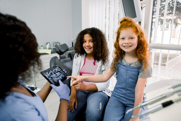 Children at dentist's office. Two multiethnic teen girls, sitting in dentistry chair, looking at camera and pointing on tablet with teeth panoramic scan together with her female african dentist