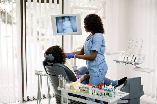 Dental care. Pretty African female doctor making examination of teeth of little patient, african american girl sitting in dentistry chair, and showing on the digital screen image of her teeth.