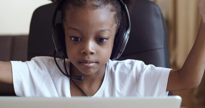 Headshot african child ethnic mixed race girl sitting at table at home wears head microphone speaks on video call online chat conference with teacher remotely, e-learning during pandemic quarantine