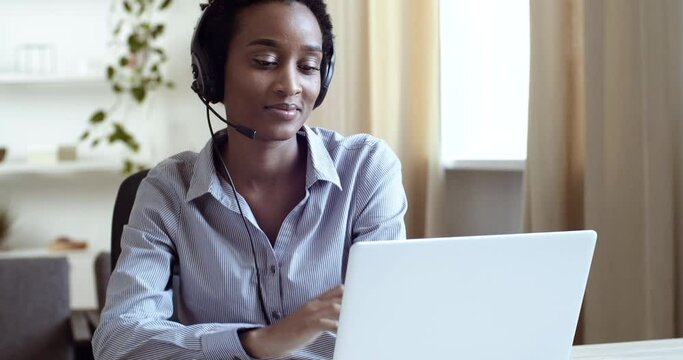 Young black girl with short hair foreign student learns language remotely listening in headphones teacher speaks with tutor on video conference watches lesson online in laptop makes notes in notebook