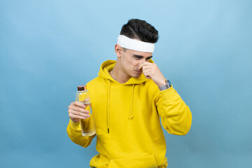 Young caucasian man wearing sportswear holding water bottle over isolated blue background smelling...
