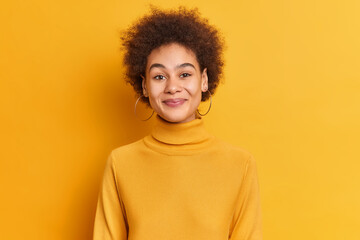 Fototapeta na wymiar Portrait of pleasant looking woman with curly hair being in good mood smiles satisfied wears casual turtleneck and earrings isolated over vivid yellow background. Satisfied African American girl