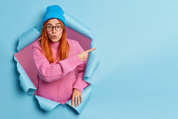 Shocked excited redhead teenage girl peeps through hole in paper points index finger on copy space...