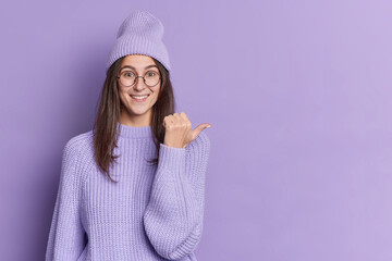 Good looking brunette young woman looks glad at camera points thumb right wears purple hat knitted...