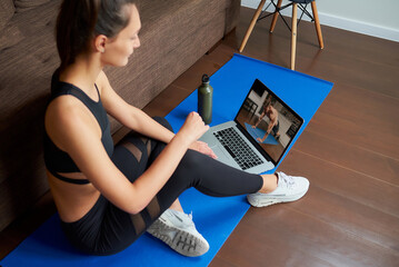 A laptop screen view over a woman's shoulder. A fit girl in a tight suit is watching a workout video on laptop in her apartment. A woman is listening to a coach during a virtual fitness class at home