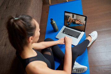 A laptop screen view over a woman's shoulder. A fit girl in a tight suit is watching a workout video on a laptop in her apartment. Woman is listening to a coach during an online fitness class at home
