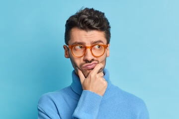 Headshot of displeased pensive adult man holds chin purses lips and looks aside tries to make decision wears spectacles and turtleneck isolated over blue background. People and thoughts concept