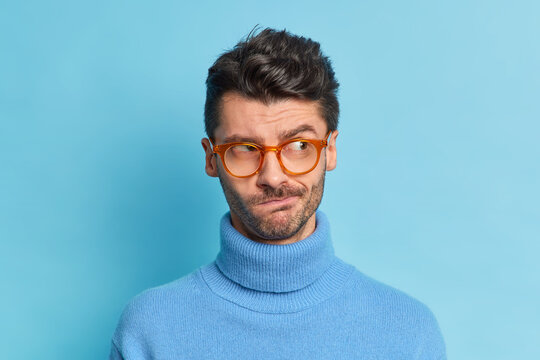 Close up shot of serious thoughtful brunet man has thick bristle purses lips wears transparent glasses and poloneck poses against blue background concentrated aside feels hesitant about something