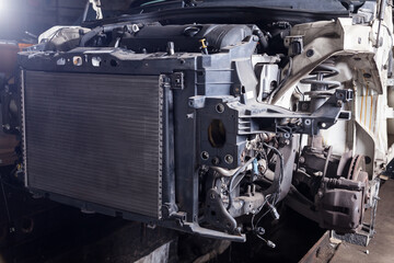 The front part of the car after an accident in a car repair service with a disassembled hood and metal grille and various parts in the garage box.