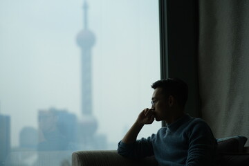 mid shot of one Asian young man sitting near window thoughtfully, blur modern landmark out of the window 