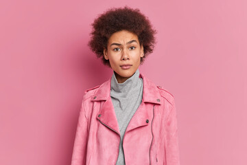 Puzzled indignant young curly African American woman frowns face raises eyebrows feels bewildered dressed in turtleneck and jacket poses against pink background. Negative human reactions concept