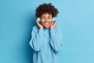 Positive curly haired woman bites lips stands with closed eyes broad smile enjoys favorite melody...
