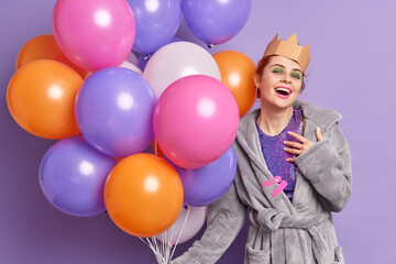 Fototapeta na wymiar Positive birthday girl feels pleased to receive congratulations from friends dressed in dressing gown handmade corona holds big bunch of multicolored balloons isolated over purple background