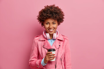 Pleased pretty young African American woman holds disposable cup of aromatic coffee wears headphones around neck dressed in stylish jacket expresses positive emotions isolated over pink background
