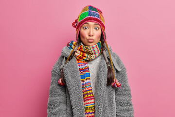 Photo of young Asian woman keeps lips rounded has surprised expression wears knitted hat and scarf dressed in grey fur coat isolated over pink background. Winter clothes face expression concept