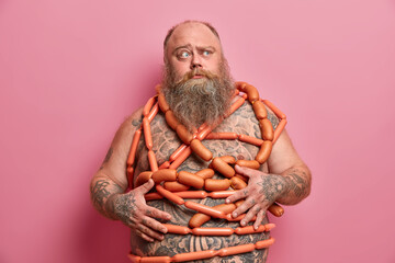 Puzzled bearded man wrapped with sausages has fat tattooed belly excess weight isolated over pink...