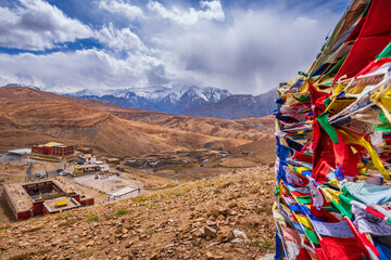 Colorful prayer flags flying in winds with Komic village nestled in  Himalayas mountains in background in Spiti valley of Himachal Pradesh, India. - obrazy, fototapety, plakaty