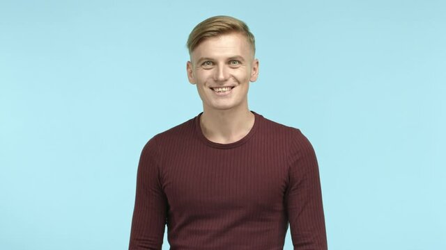 Handsome caucasian man with blond hair looking excited at camera and saying yes, standing amazed over blue background