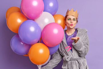 Fototapeta na wymiar Romantic beautiful woman blows air kiss dressed in domestic clothing has bright vivid makeup holds big bunch of balloons expresses love to you poses over purple background. Party holiday celebration