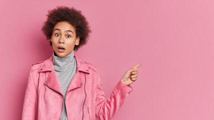 Obraz na płótnie Canvas Emotive female teenager with Afro hair keeps jaw dropped from shock demonstrates amazing promo wears fashionable jacket points right on blank space demonstrates big shopping sale. Advertisement