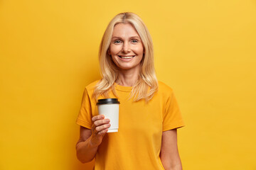 Horizontal shot of blonde European woman with pleasant smile minimal makeup holds disposable cup of coffee dressed in casual t shirt isolated over yellow studio wall. People age lifestyle concept