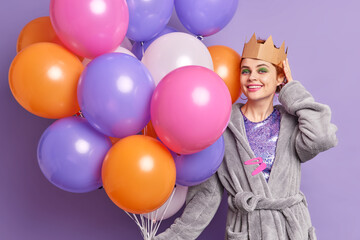 Positive woman with bright makeup wears paper crown and dressing gown organizes party at home holds many helium colorful balloons smiles pleasantly poses with holiday attributes against purple wall