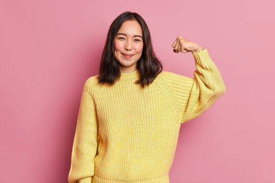 Horizontal shot of pretty Asian woman raises arms shows muscles being strong person has power dressed in casual yellow jumper poses against pink background. Look at my biceps after training.