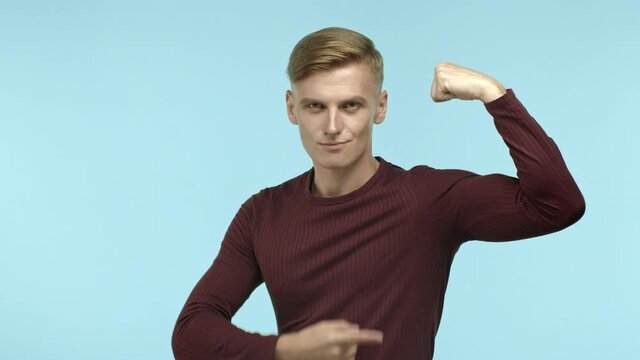 Confident and sassy blond fit man flexing biceps, pointing at strong muscle hand and raising eyebrow with smug face, standing over blue background. Concept of workout and healthy lifestyle