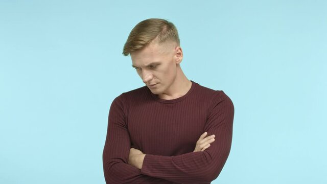 Handsome blond man looking troubled and complicated, cross arms on chest, looking at camera serious and thoughtful, making difficult decision, blue background