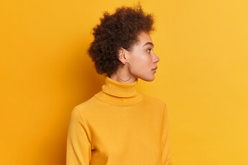 Fototapeta na wymiar Studio shot of curly woman stands sideways against yellow background turns head aside has serious expression dark curly hair dressed in turtleneck wears round earrings notices something on right