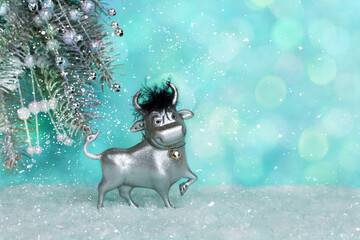 A toy bull on a blue background stands in the snow against the background of a Christmas tree. The symbol of the new 2021. Good New Year mood. Place for an inscription. The basis for the postcard.