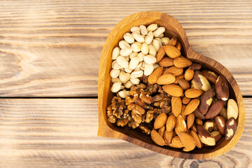 Natural nutritional blend of various nuts in a wooden plate in the shape of a heart symbol on a brown wooden table. A mixture of walnuts, brazil nuts, peanuts and almonds. Healthy natural food concept