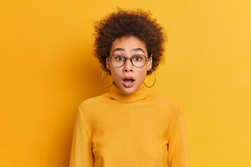 Fototapeta na wymiar Portrait of shocked curly woman gazes at camera with opened mouth reacts on amazing news wears transparent glasses earrings turtleneck. Speechless impressed Afro American teenager keeps jaw dropped