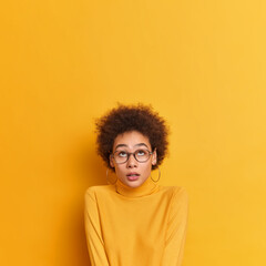 Obraz na płótnie Canvas Omg unbelievable thing above. Surprised pretty curly African American girl concentrated above holds breath from wonder wears transparent glasses casual poloneck isolated over yellow background.