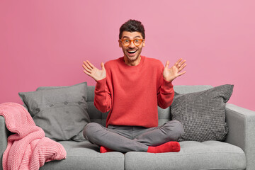Joyful man raises palms reacts on awesome news sits crossed legs on comfortable sofa wears casual...