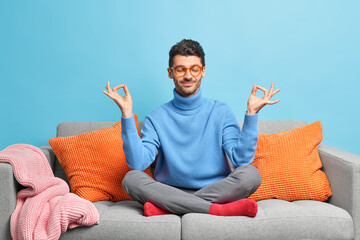 Calm relaxed man sits in lotus pose meditates at home during quarantine poses on comfortable sofa...