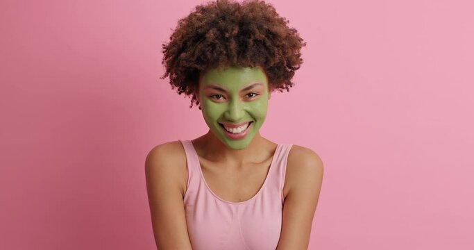 Skin beauty concept. Positive young Afro American woman looks gladfully around undergoes facial treatments smiles broadly models against pink background. Rejuvenation and cosmetology concept