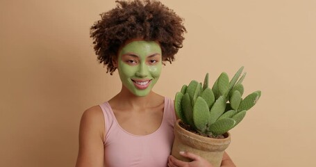 Beautiful African American woman smiles gently has perfect skin applies green nourishing mask for face care dressed casually holds potted cactus isolated over brown background. Anti aging procedure