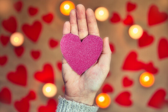 Female Hand Holding A Glitter Pink Heart With Romantic Candle And Red Hearts Background For Valentine's Day Or Romantic Concept