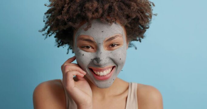Marry lovely woman with curly bushy hair touches camera with index finger smiles broadly applies clay facial mask shows bare shoulders puts facial mask enjoys beauty treatments poses indoor.