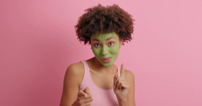 Positive dark skinned woman dances carefree raises clenched fists and smiles broadly indicates at you has fun at home undergoes beauty procedures applies green beauty mask isolated on pink wall